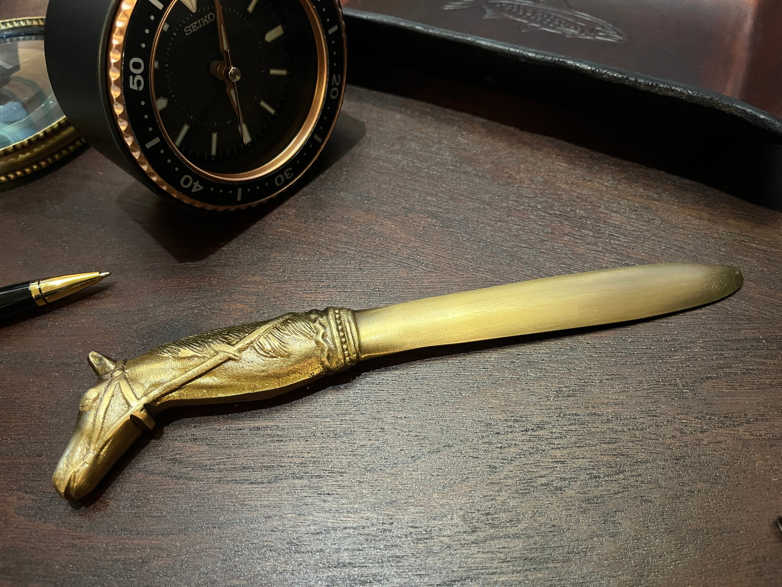 Brass letter opener shaped like a horse head on a wooden surface with a clock in the background.