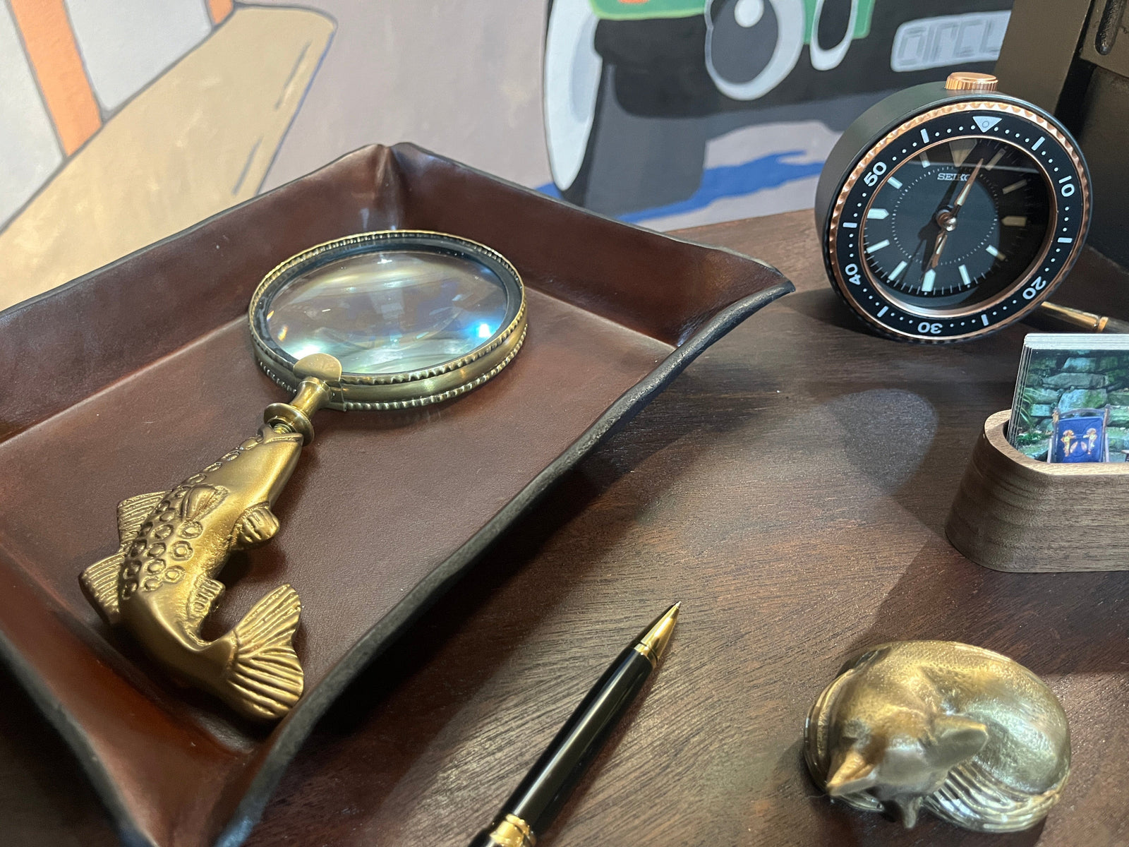  Fish shaped magnifying glass, compass, and other items on a wooden surface with a colorful abstract painting in the background.