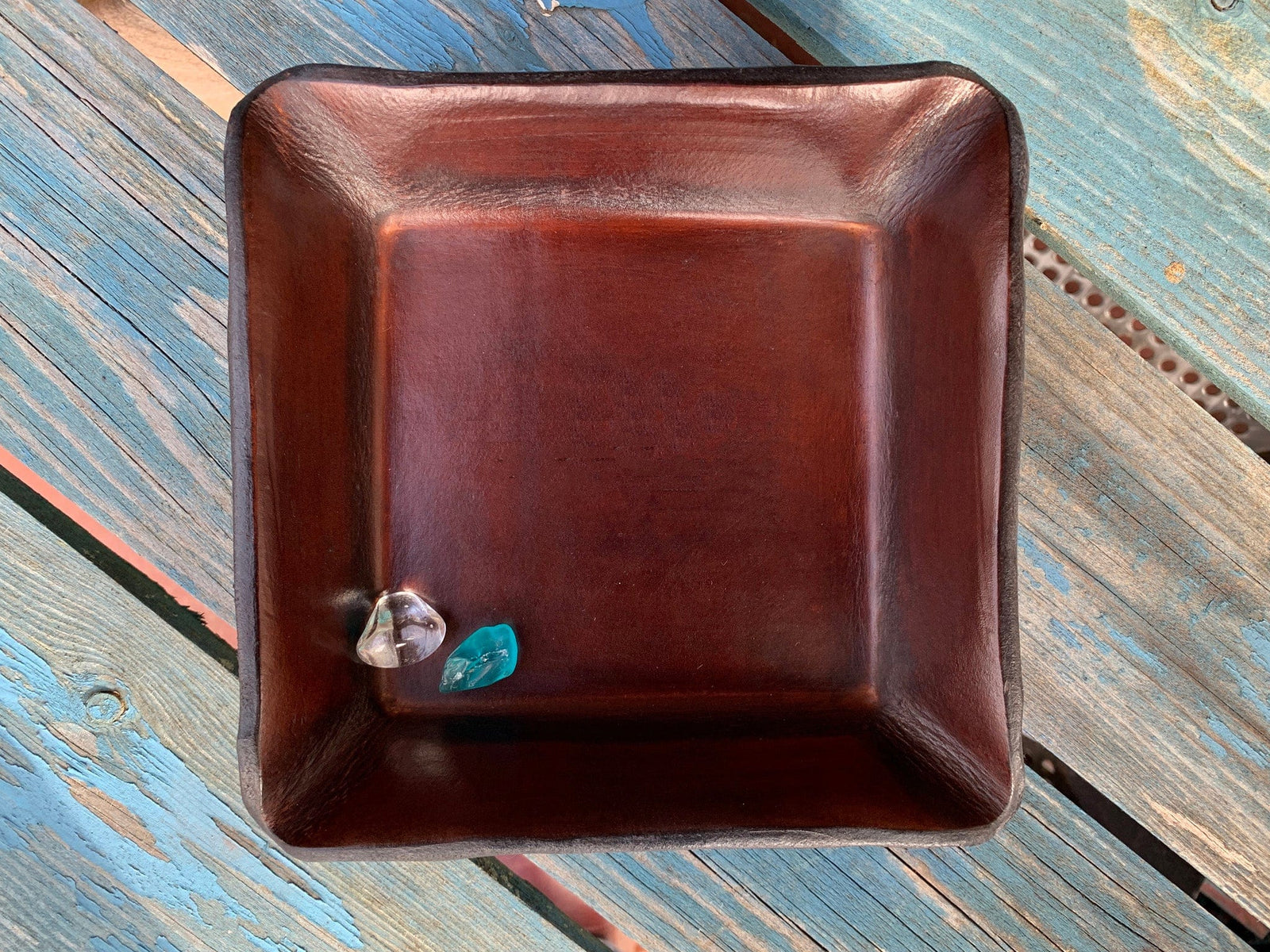 Square, brown leather tray shown with sea glass inside on a blue, wooden surface