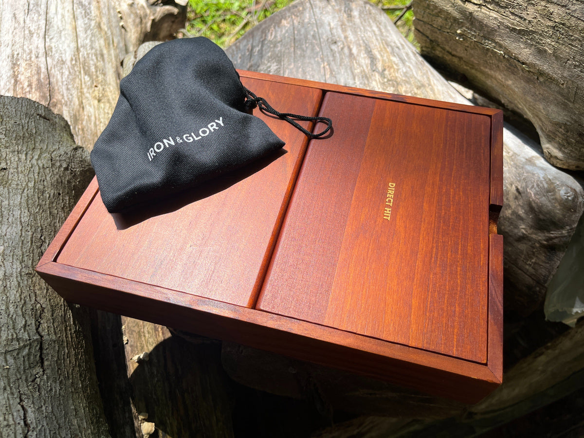 Wooden game box with a black bag labeled &#39;Iron &amp; Glory&#39; on top, set against a natural background of rocks and trees.